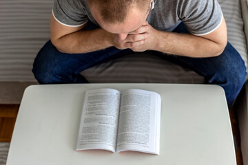 Young man sitting in the lounge and reading a book. Adult Caucasian man sitting on sofa bed at home reading a book, study, learning alone during the quarantine lockdown. Education, lifestyle concept.
