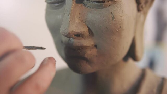 Close up of clay Buddha's statue face, artist works with details using spatula