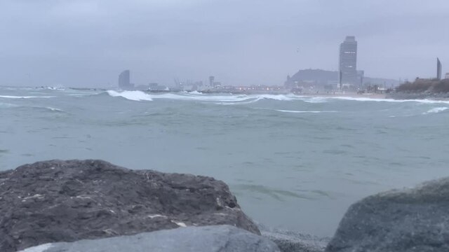 Big waves in storm on Barceloneta beach with rocks in the foreground and Wella hotel in the background