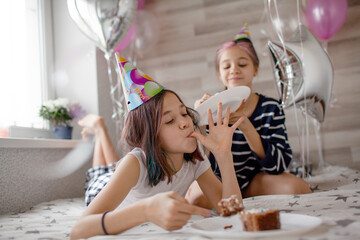 Two preteen sisters twins celebrate birthday day at the morning, active kids dressed in pajamas have fun in the bed with balloons. Children eat cake in the bed