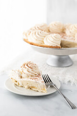 Close up of a slice of banana cream pie with the full pie in behind against a bright sunny window.