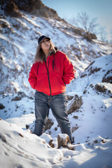 A beautiful girl in a red down jacket, jeans and a black baseball cap stands in the middle of the snow dunes somewhere in the mountains and looks into the distance.