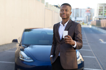 Portrait of young handsome African American businessman or sales manager, standing with a card key from a new electric car outdoors at dealership. Concept of eco-friendly and modern cars