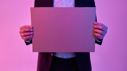 Cropped image of young attractive businessman in black suit and white shirt holding cardboard in hands with empty space for text isolated on neon pink background.
