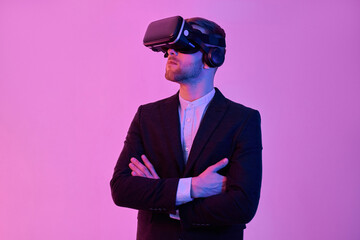 Obraz na płótnie Canvas Young attractive businessman in a black suit and white shirt uses virtual reality glasses, stands with his arms crossed, isolated on a neon pink background. Future technologies.