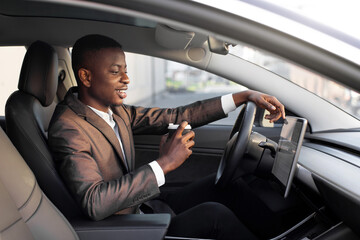 Young excited African businessman, having his lunch break to drink take away coffee, while sitting in modern electric car with self-steering system, and looking at car touchscreen display