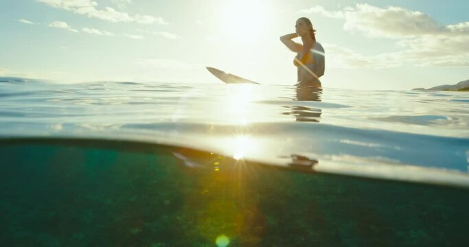 Beautiful female surfer sitting on surfboard in ocean at sunset waiting for the next wave. Slow motion shot on RED cinema camera.
