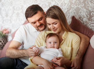 Fototapeta na wymiar Beautiful young parents sit on the sofa with their newborn daughter in their arms in a light room