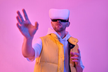 Young attractive man in casual clothes, uses virtual reality glasses, trying to touch something with his hand, isolated on neon pink background. Future technologies.