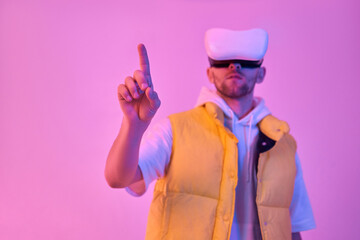Young attractive man in casual clothes, uses virtual reality glasses, trying to press the virtual button from scratch isolated on neon pink background. Future technologies.