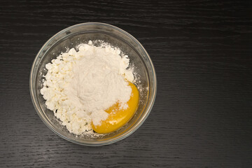 Grainy cottage cheese, egg, flour in a glass bowl on a black table close-up with copy space, top view