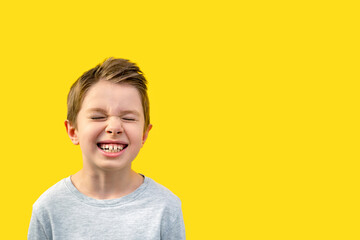 Portrait of a handsome boy closed and screwed up his eyes and laughs with his mouth wide open, a child in a gray shirt smile, positive emotions, childhood, school age. Yellow background, copy space - obrazy, fototapety, plakaty