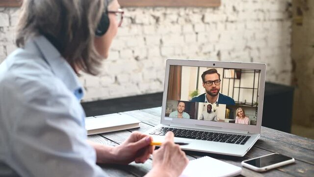 Mature gray-haired businesswoman in headset having virtual meeting with the multiracial team, online business video call via laptop from home office. Senior woman looking at the screen and talking