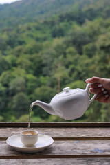 A Woman hand pouring a tea into the cup on the wooden table which has the background of mountain and forest