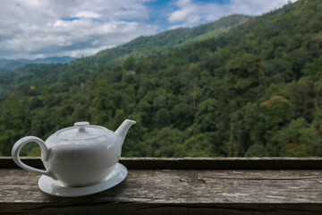 The white teakettle on the wood table with the view of mountain and forest as the background