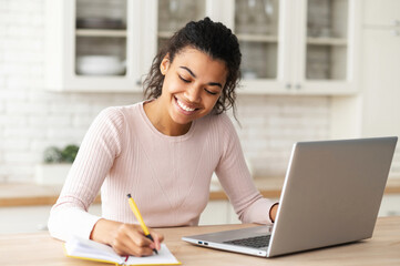 Positive African American female student smiling sitting at the table with laptop in the kitchen, studying from home and taking notes, writing to the notebook, watching online webinar, taking courses