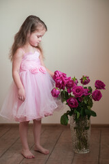 A cute little girl with long hair in a beautiful pink dress chooses a flower from a vase with roses that have faded a little