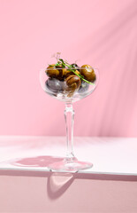 Olive Kalamata in cocktail glass. Black and green olives food on white table with pink wall. Day sunlight with hard shadow of fern palm shadow.