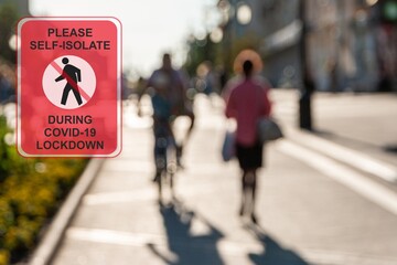 Concept ''Please self-isolate during COVID-19 lockdown'' road sign against a defocused bicyclist and woman in the city