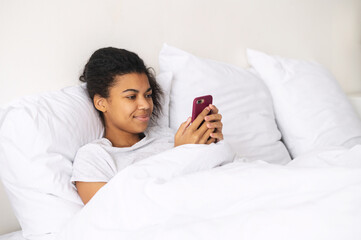 Happy young mixed-race woman with curly hair covered with blanket staying at home in warm bed, chatting with friends on mobile phone, flirting, scrolling social media, writing good morning message