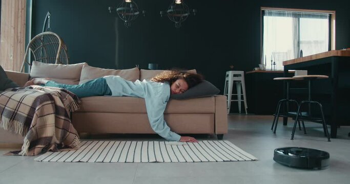 Carefree young African American housewife woman tired sleeping on couch at home, robotic vacuum cleaner sweeping floor.
