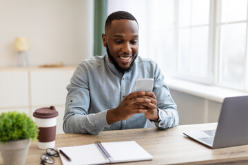 Happy African Businessman Using Phone App Sitting At Workplace Indoors