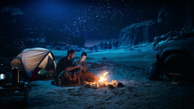 Happy Couple Nature Camping in the Canyon at Night, Use Digital Tablet Computer, Sitting by Campfire. Two Traveling people Have Fun, Smile, Post on Social Media, Watch Funny Videos on Internet