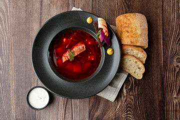 Ukrainian traditional borshch. Beetroot and cabbage soup with meat served with sour cream and sliced bread by side. Isolated dish in black plate on wooden background and grey linen napkin. Top view.