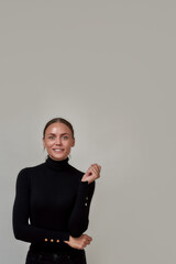 Elegant caucasian young woman wearing black turtleneck smiling at camera, holding arm, standing isolated over gray background