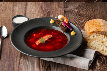 Ukrainian traditional borshch. Beetroot and cabbage soup with meat served with sour cream and sliced bread by side. Isolated dish in black plate on wooden background and grey linen napkin.