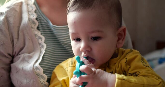 A young mother sits on the bed with a small child in her arms and controls the game with a teether and other tactile toys. Maternity, motherhood and family concept. slow motion