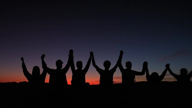 Silhouette showing group of people holding hands together as a team in front of colorful sunset. Close up pan shot.