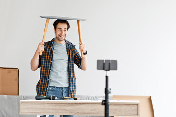 Cheerful millennial male with wireless headphones has fun with ready-made chair and raises it above his head