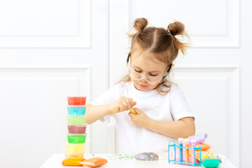 Adorible child in a white T-shirt with two tails sits at the table in protective glasses and doing experiments, making fluffy slime from multicolored ingredients. Cool kid studying chemistry science