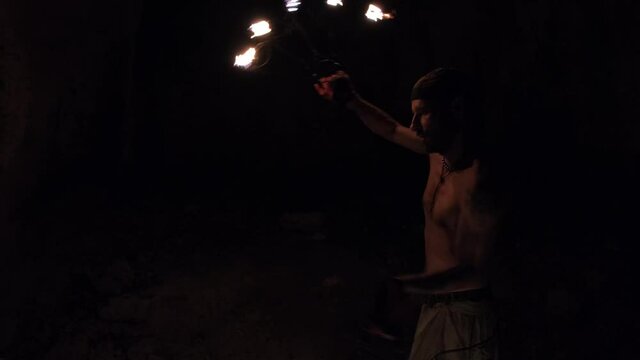 A young male performer dances with a fire fan. Exciting fire show in a dark cave.