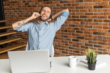 Smiling man with headset sitting in front of desk with a laptop and talking on his break, has a...