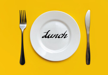 A white plate on which the word Lunch is written standing on a yellow tablecloth . The concept of a balanced diet, ration and medical fasting. Top view, white background, copy space.