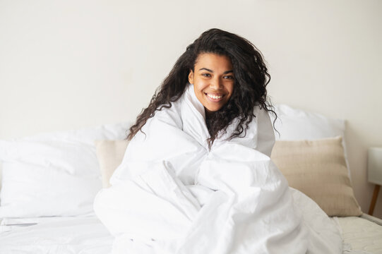 Young mixed-race woman with curly hair covered in a cozy white soft blanket staying at home in the warm bed, looking at the camera with a toothy smile, feeling happy and blessed, good morning concept