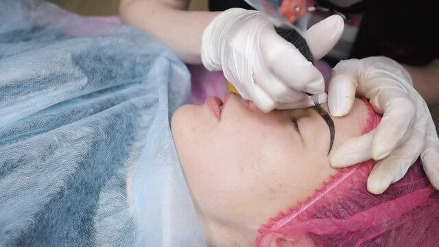 Beautician makes permanent make up correction of the shape of the eyebrows to a young beautiful girl. Microblading, eyebrow tattooing in a beauty salon. Profile view