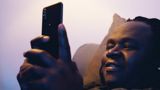 Young african american man browsing social media on his smartphone with a smile while relaxing on the couch. High quality 4k footage