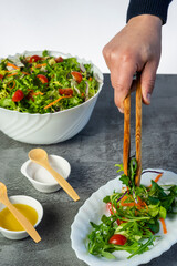 Man's hand serving salad of lettuce, tomatoes, rugula, various green vegetables with wooden tongs to an oval dish. White bowl full of salad bowl full of salad and small plates with salad dressing. 