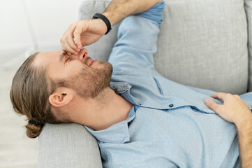 Fototapeta premium Attractive man with light brown hair pulled back, beard, lying on the grey cozy couch, with pleasure smile, keeping arm on the face, remembering this fantastic day, got long-awaited job, after date