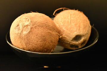 Two ripe organic coconuts on a black ceramic plate, close-up, isolated on black.