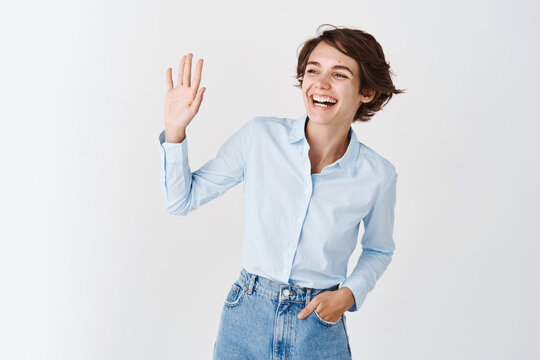 Young candid woman laughing and smiling aside, waiving hand at empty space to say hi, greeting person and make hello gesture, standing on white background