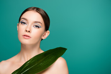 young woman with creative makeup looking at camera near wet leaf isolated on green