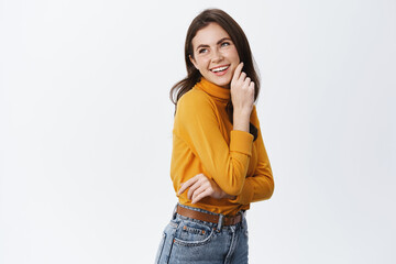 Beautiful girl standing half-turned and looking left with cheerful smile, touching cheek and staring happy, wearing casual clothes, white background