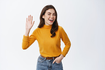 Hey hello. Smiling friendly girl saying hi, waving hand and looking happy at camera, greeting and introduce herself, standing against white background