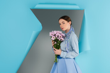 trendy woman looking at camera while holding pink flowers near hole in blue paper on grey background