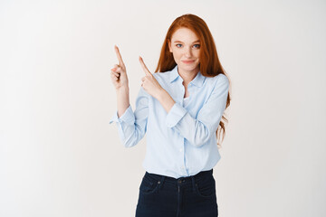 Young female student with ginger hair and blue eyes showing company logo, pointing fingers upper left corner and smiling at camera, white background