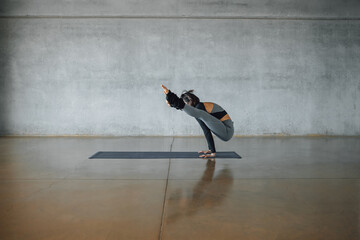 Full-length female yoga instructor, doing difficult Firefly pose on a mat on the floor of a urban...